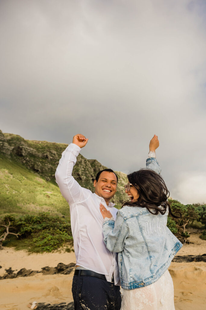 Couple cheers after just getting married on the beach in Oahu, Hawaii with mountains in the backrgound