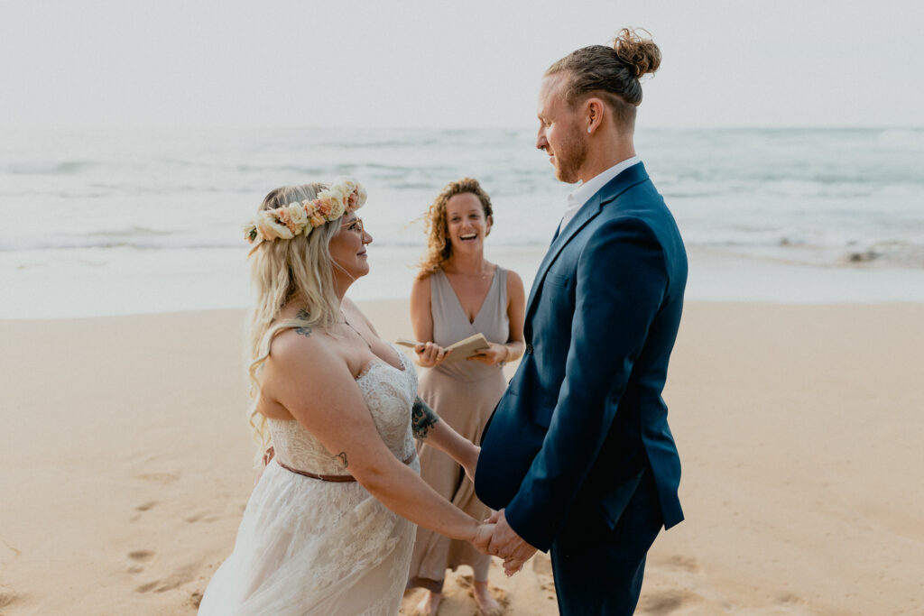 Couple stands on the beach on Oahu, holding hands on their wedding day with their wedding officiant the background.