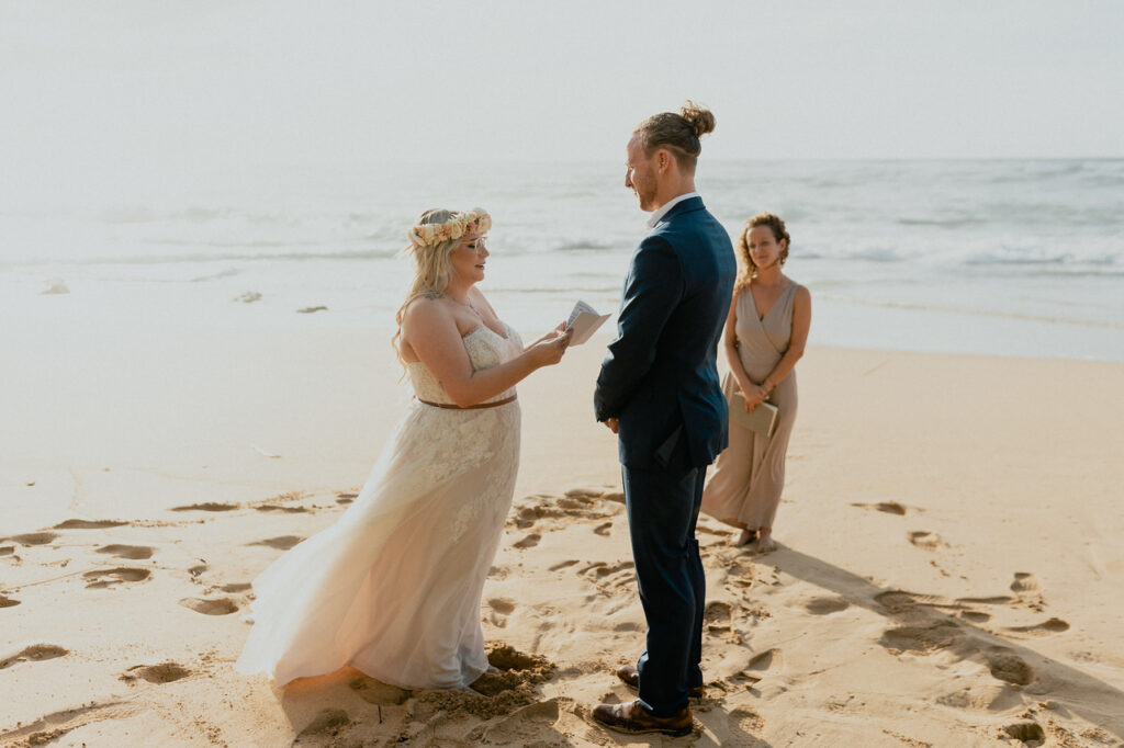 Couple stands on a beach in Oahu, Hawaii. As they read their wedding vows to one another their wedding officiant stands in the background. 